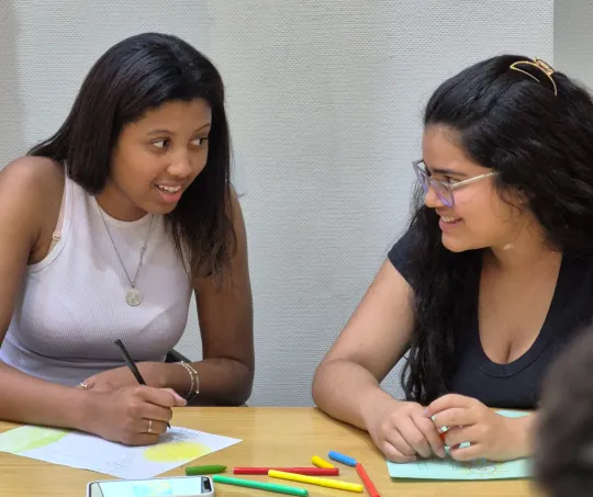 Two young women sitting at a table, smiling and engaged in drawing with colorful markers and paper.
