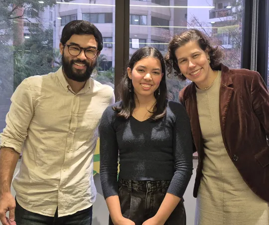 Three people smiling and posing indoors in front of a large window with city buildings outside.