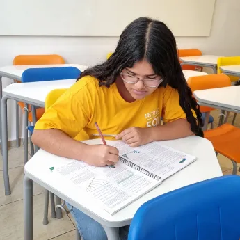 Student with glasses and braided hair wearing a yellow shirt, writing in a notebook at a desk in a classroom with colorful chairs.