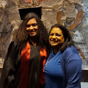 Young woman in a graduation cap and gown standing next to an older woman in a blue top, both smiling.