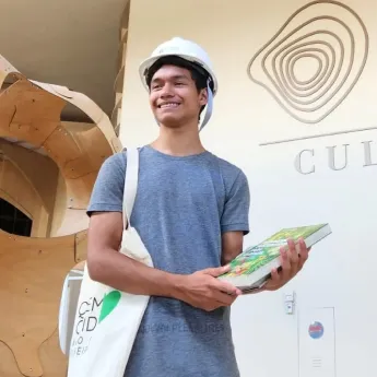 Smiling young man wearing a white safety helmet and gray t-shirt holding a book and a tote bag standing in front of a building wall with an abstract design.