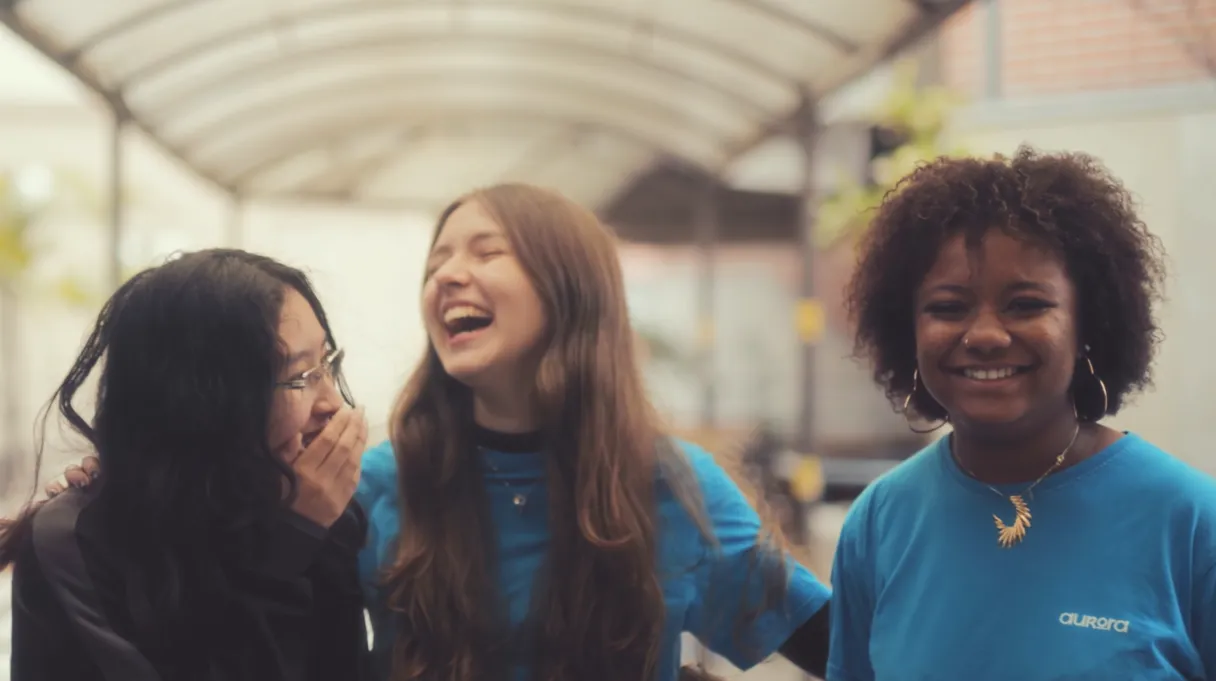 Three young women smiling and laughing together under a covered outdoor walkway.