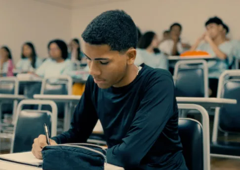 Student focused on writing in a classroom with peers blurred in the background.