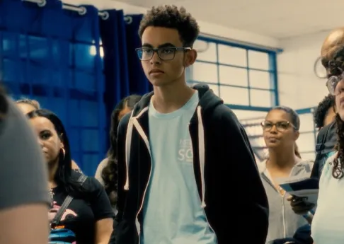 Young man with glasses and curly hair wearing a light blue shirt and black hoodie standing among a group of people indoors.