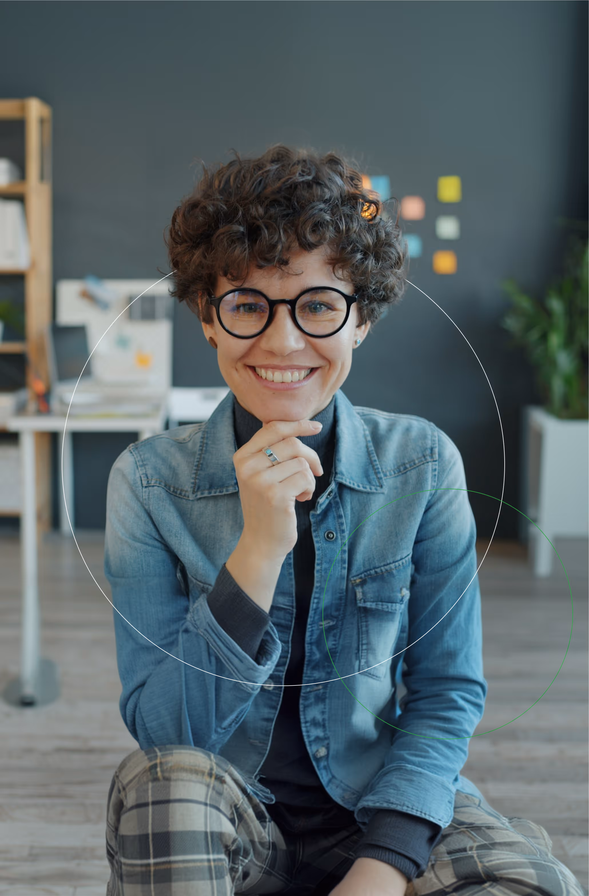 Smiling person with curly hair, black round glasses, denim jacket, and plaid pants sitting indoors with a hand near their chin.