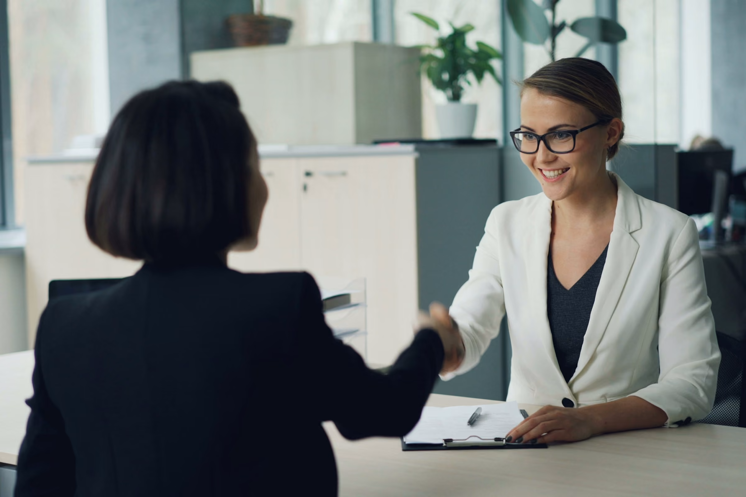 Two professional women shaking hands across a desk in a modern office setting.