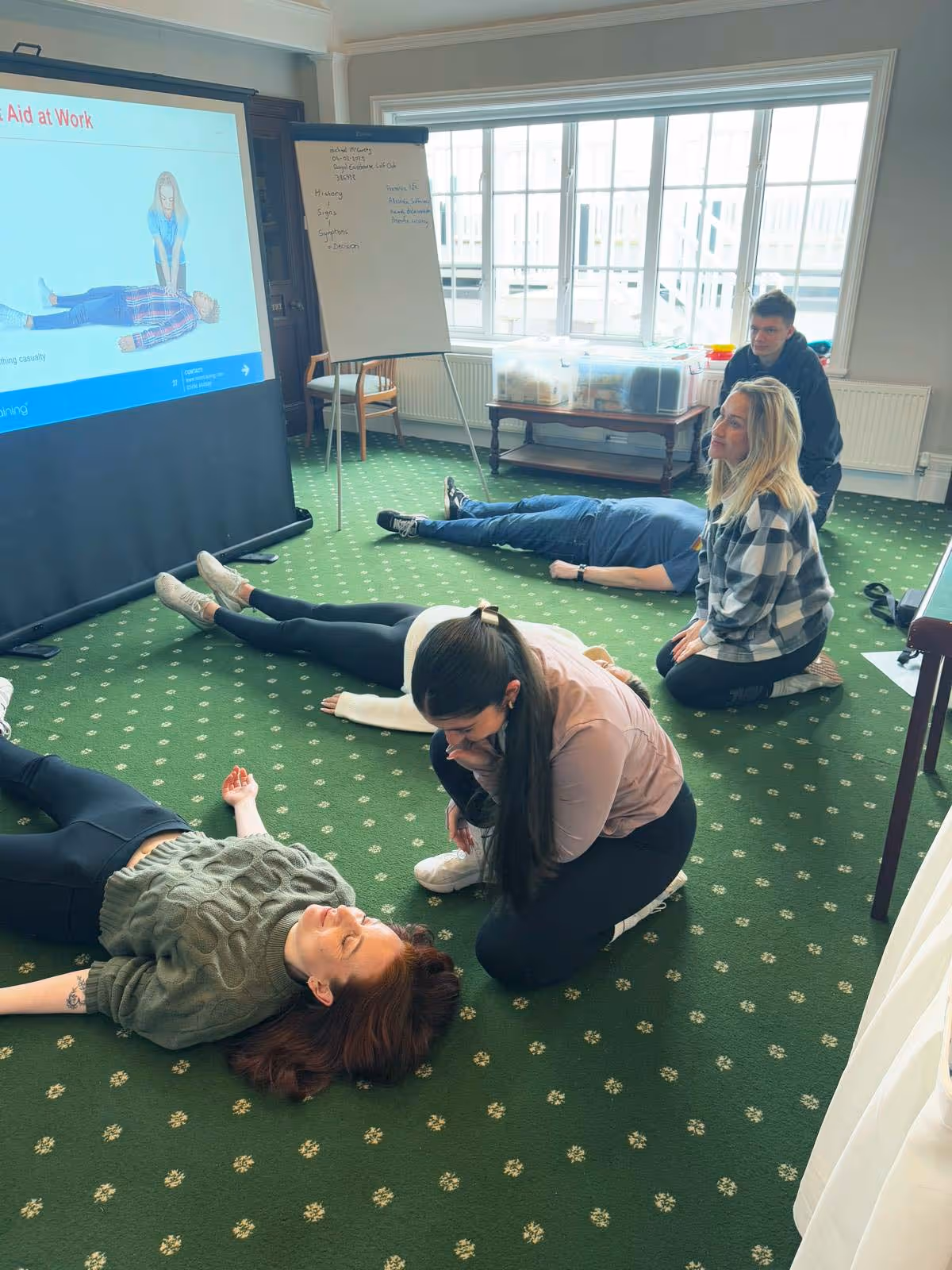 People practice first aid, performing recovery position exercises in a classroom setting on green carpet.