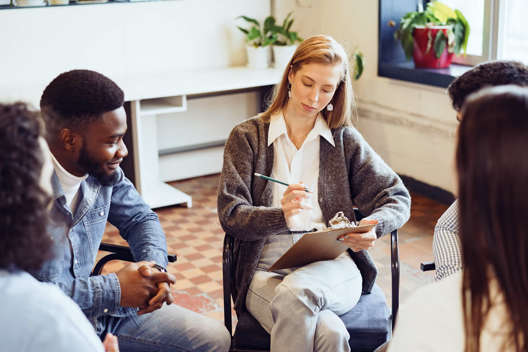 A group of people sits in a circle, while a woman writes notes on a clipboard.