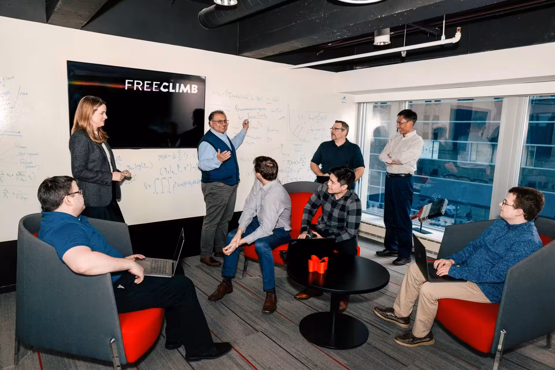 A group of seven researchers gather closely together, looking intently at a laptop screen. They are in an office with writing and diagrams on a whiteboard in the background. They appear engaged and focused on the content displayed.