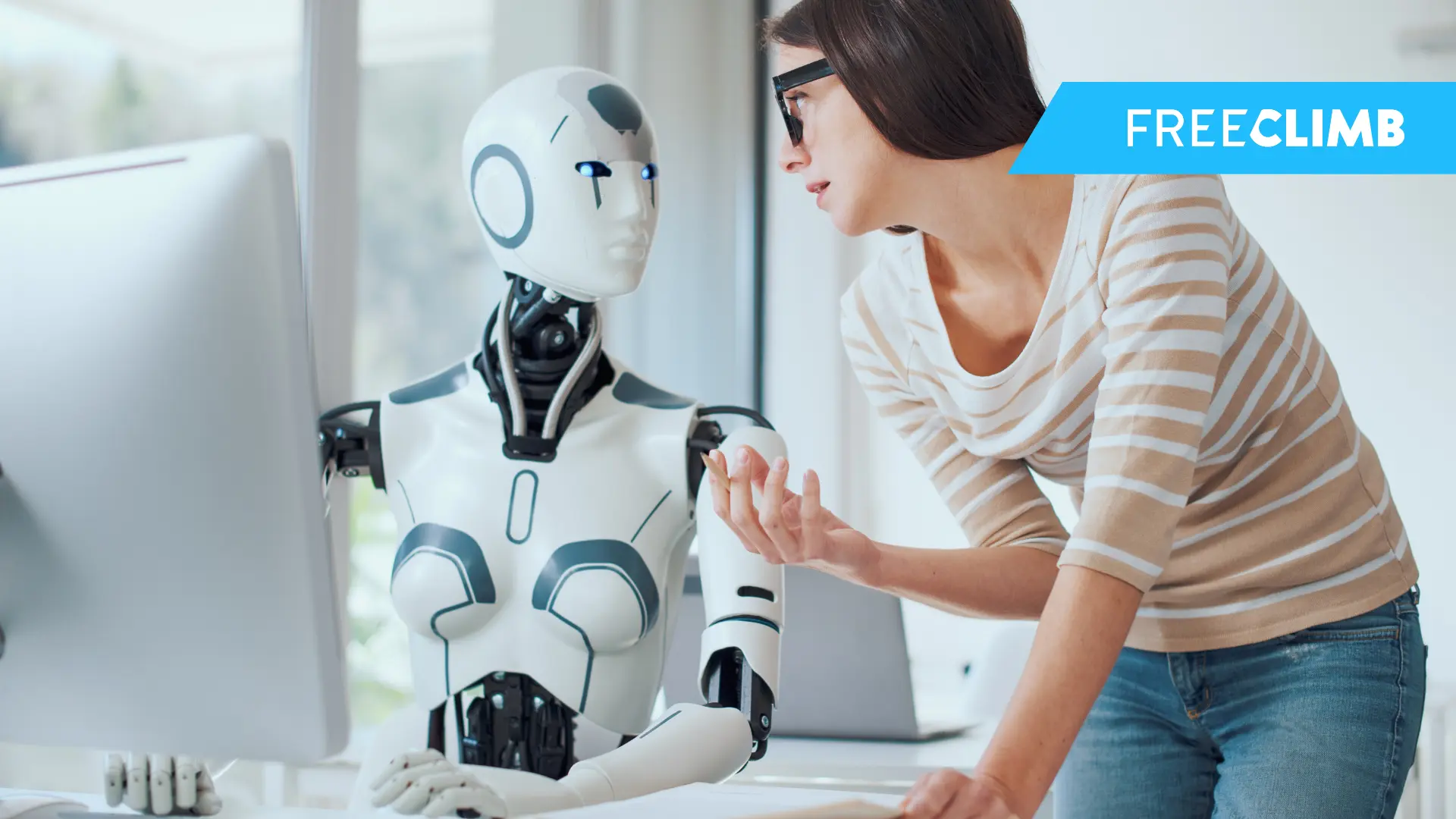 A robot sits at a computer while a woman stands beside it, speaking over its shoulder.