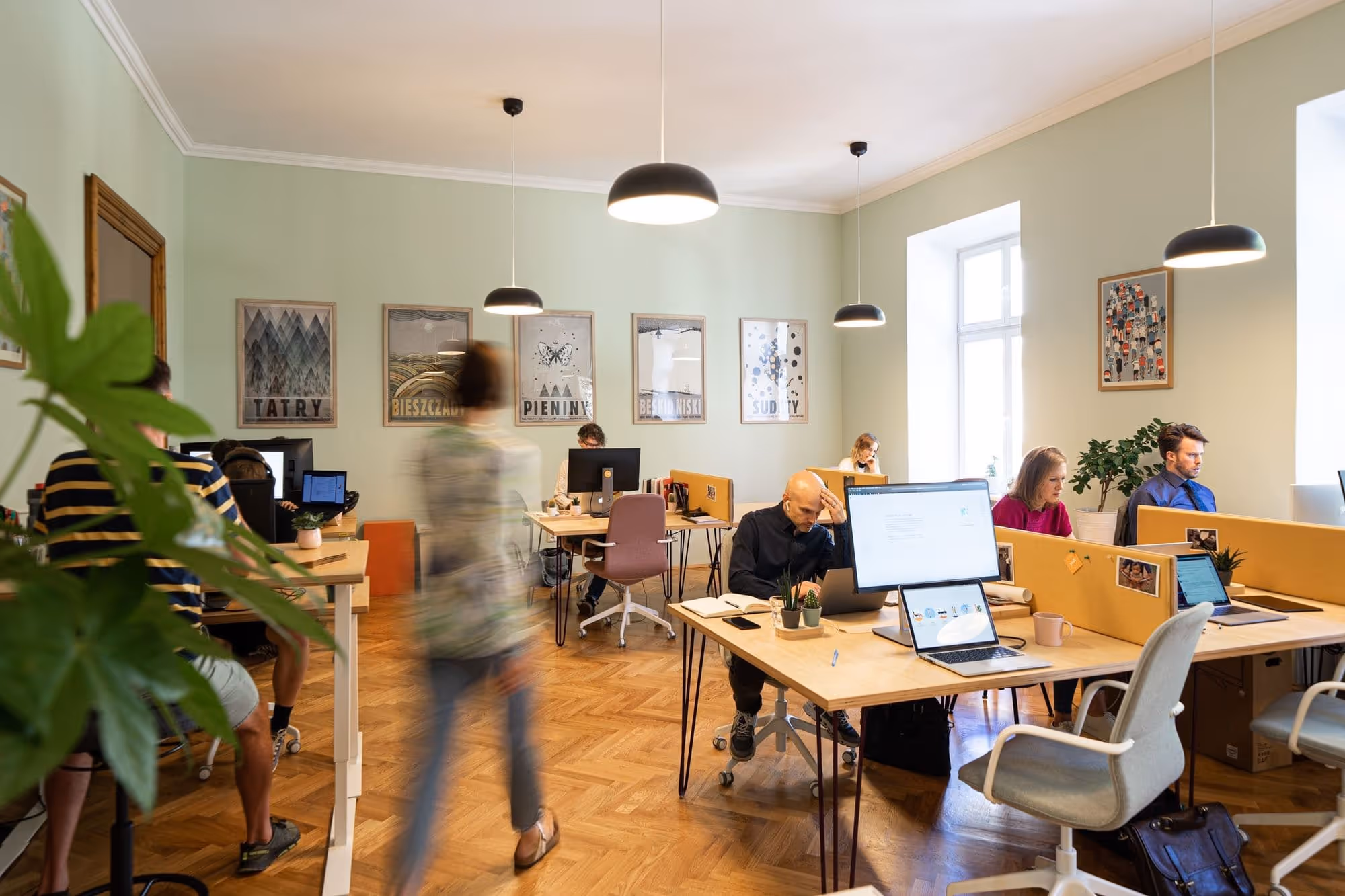 Modernes Büro mit mehreren Menschen, die an Schreibtischen mit Computern arbeiten, warm beleuchtet und mit Pflanzen dekoriert.