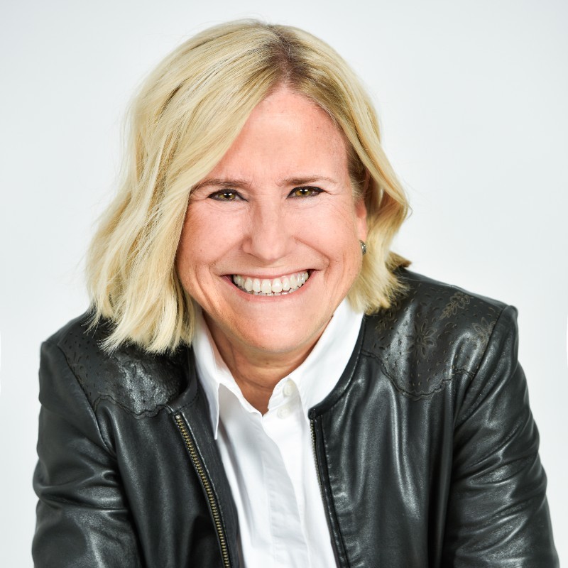 Smiling middle-aged woman with short blonde hair wearing a black leather jacket and white shirt.