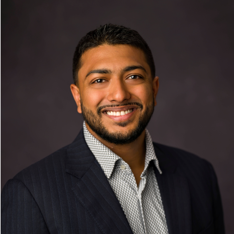 Smiling man with short black hair and beard wearing a patterned shirt and dark blazer.