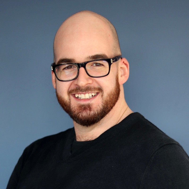 Smiling bald man with beard and glasses wearing a black shirt against a plain blue background.