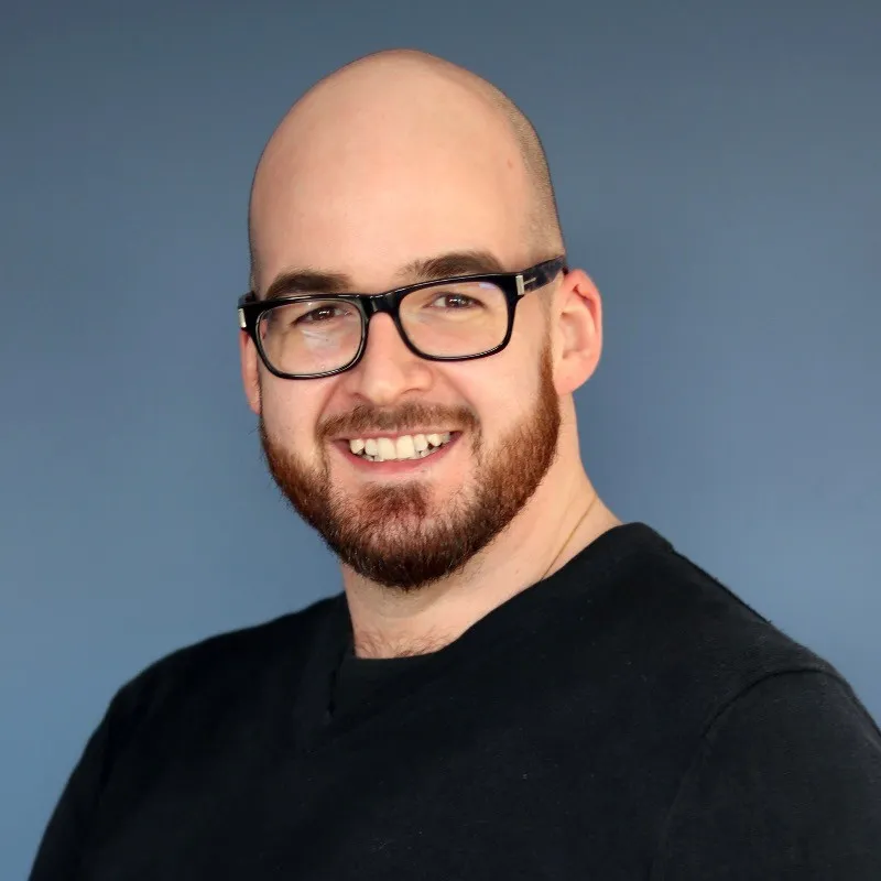 Smiling bald man with beard and glasses wearing a black shirt against a plain blue background.