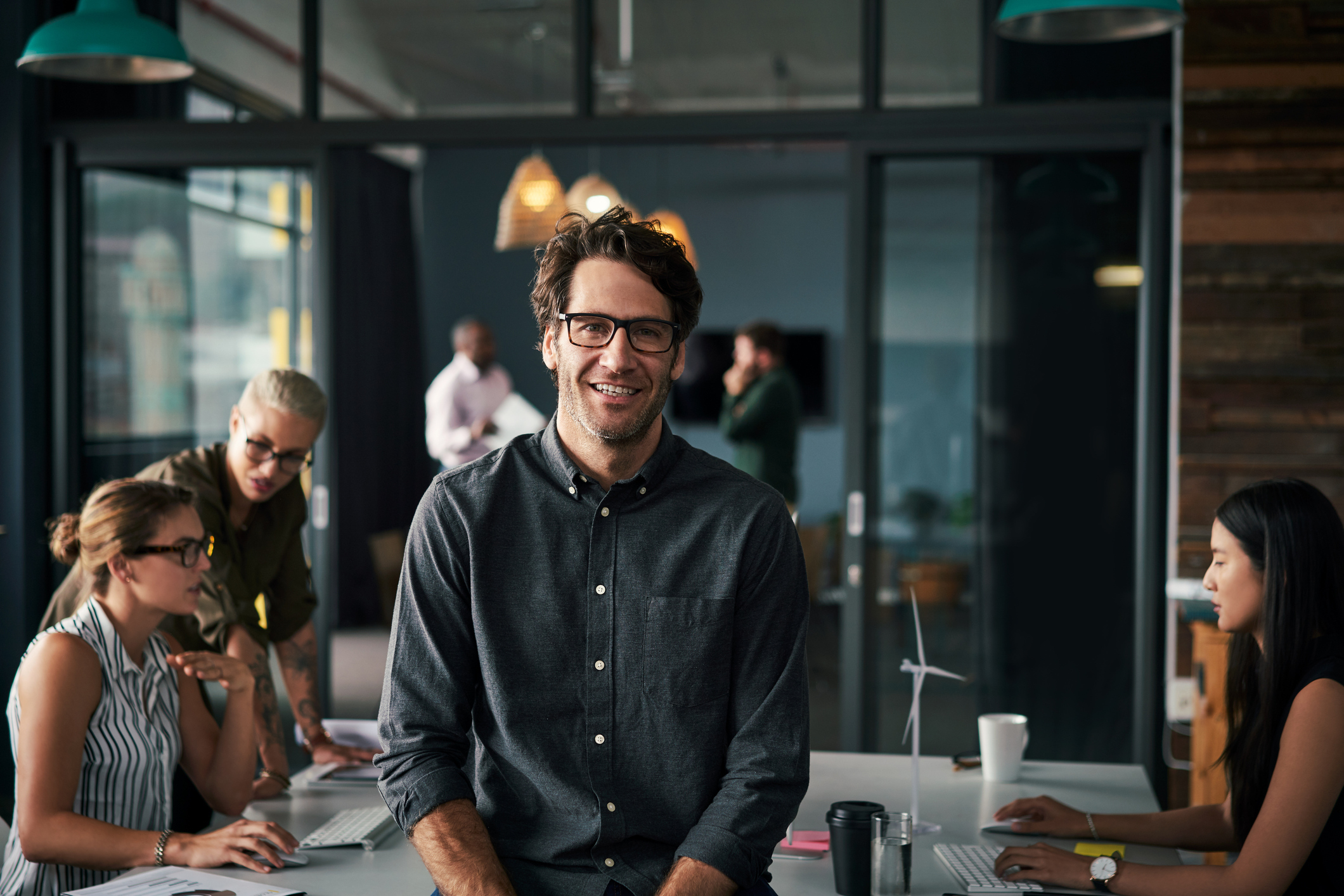 Man smiling and sitting at a table in a modern office with colleagues working around him.