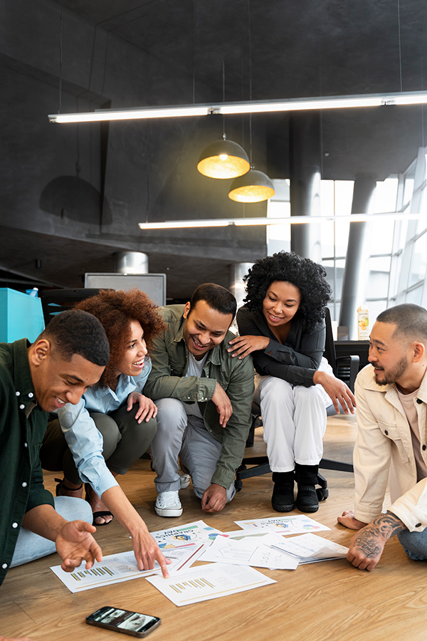 Five diverse coworkers sitting on the floor in an office, reviewing charts and documents together.