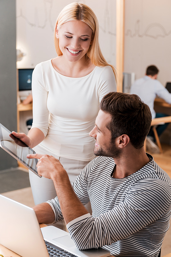 Smiling woman shows tablet to seated man who points at screen in office setting.