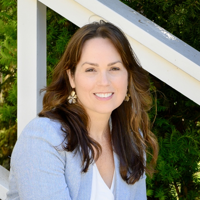 Smiling woman with long brown hair wearing a light gray blazer and white top outdoors.