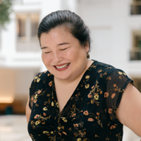 Smiling woman with dark hair wearing a black floral blouse in a bright indoor space.