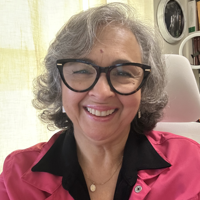 Smiling woman with glasses and gray curly hair wearing a pink shirt and black collar.