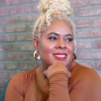 Smiling woman with blonde hair in a bun, wearing gold hoop earrings and a brown top, against a brick wall.