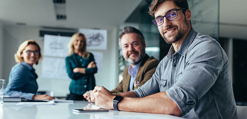 Four professionals in a meeting room, one smiling man in front with glasses and a smartwatch.