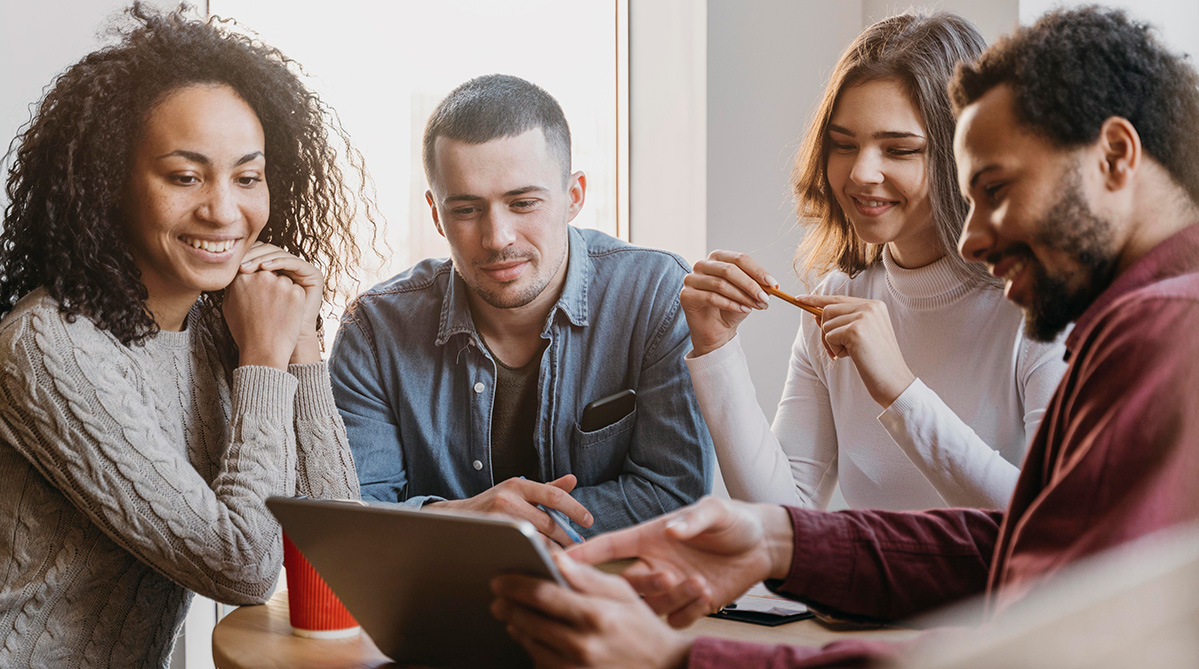 Four diverse coworkers smiling and discussing something on a tablet around a table in a bright room.