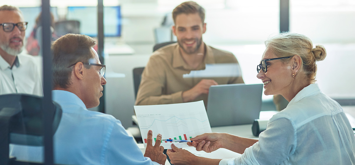 Four professionals discuss a report with charts in an office meeting, smiling and engaged.