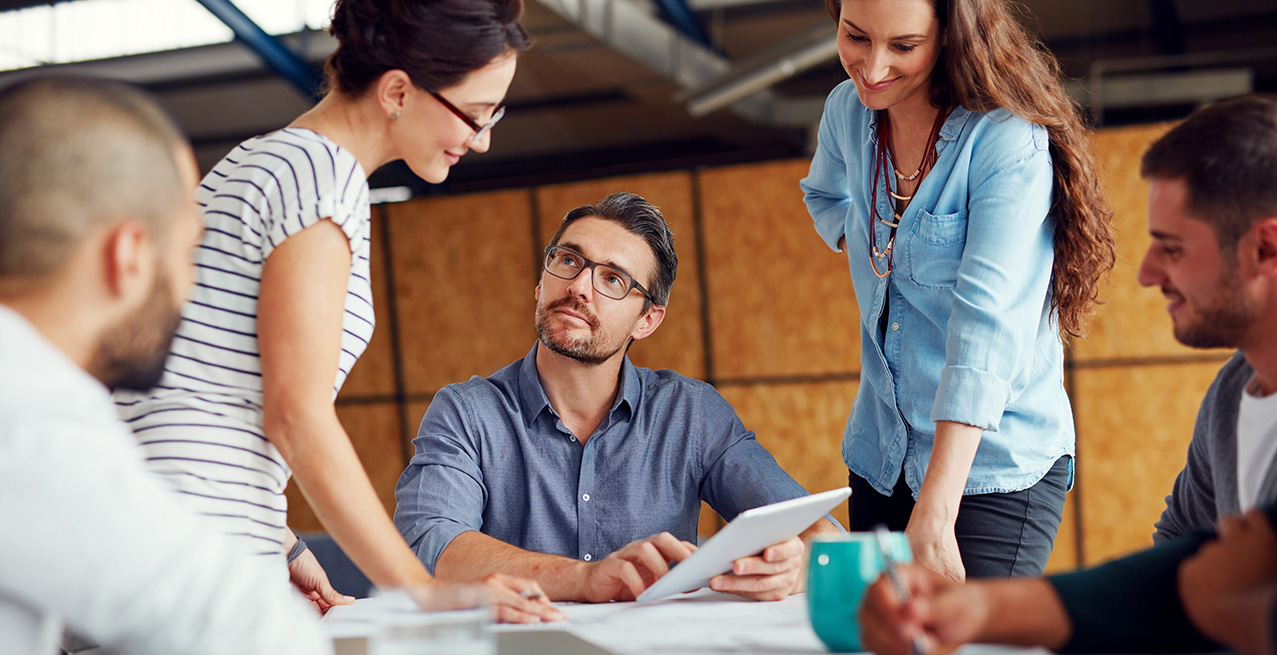 Five diverse coworkers collaborating around a table, one holding a tablet, in a modern office setting.