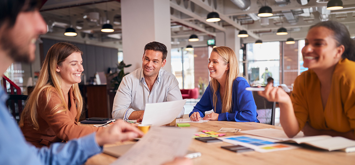 Four colleagues smiling and discussing documents around a table in a modern office.
