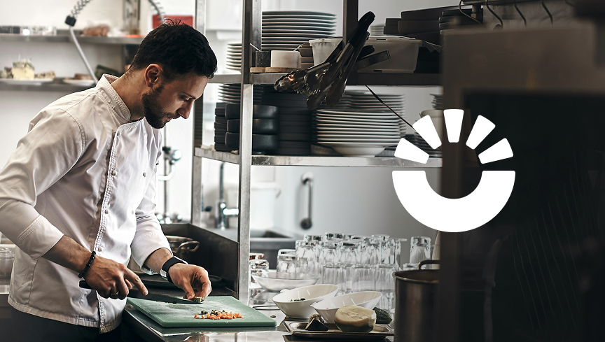 Chef in white coat finely chopping ingredients on a green cutting board in a professional kitchen.