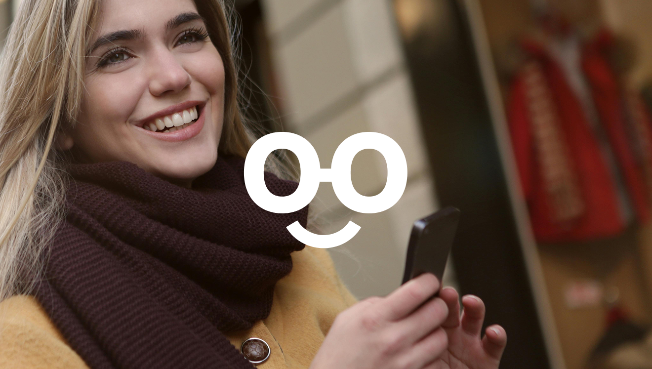 Smiling woman wearing a brown scarf and yellow coat holding a smartphone outdoors.