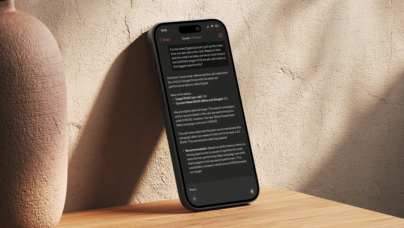 Smartphone displaying a chat conversation about ad campaign performance, resting on a wooden surface next to a large textured vase with a beige wall background.