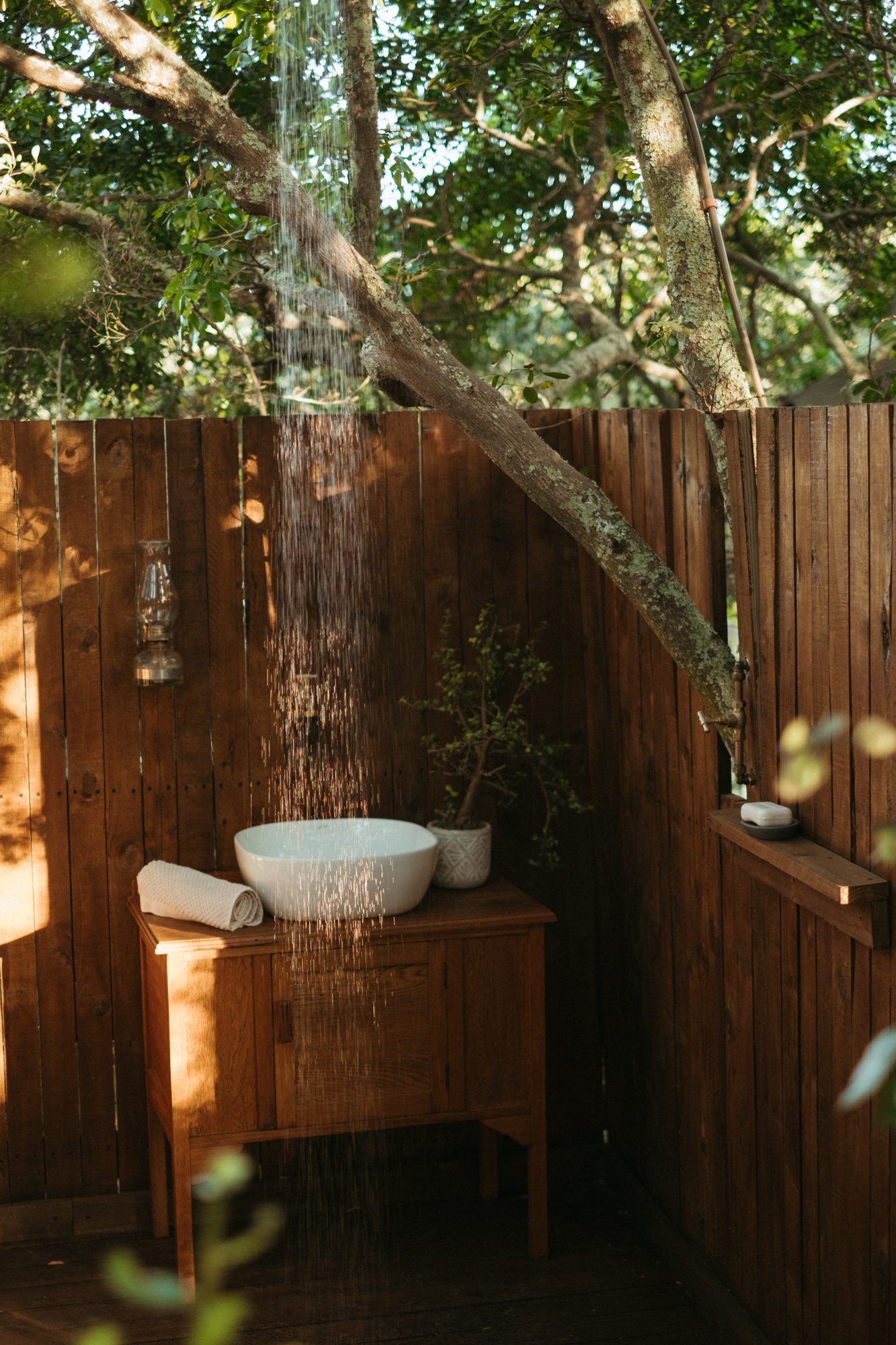 An open-air shower falling beside a basin on a timber stand, under the trees