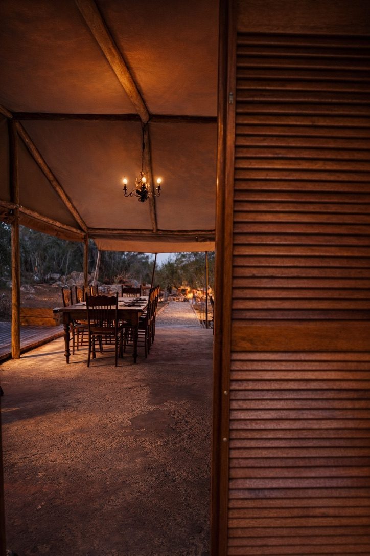 The mess tent table lit by a chandelier at dusk, seen past a timber shutter