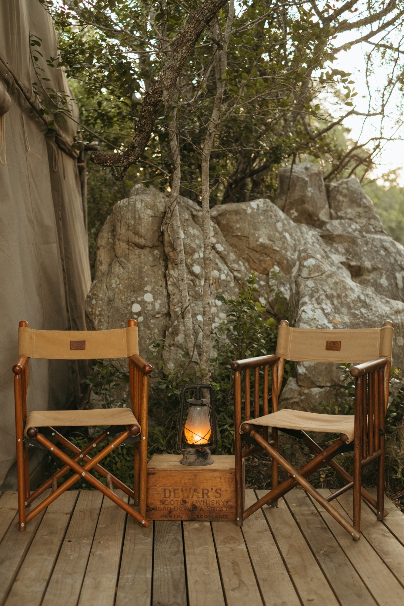 Two canvas director's chairs and a lantern on a crate on a tent deck, rocks behind