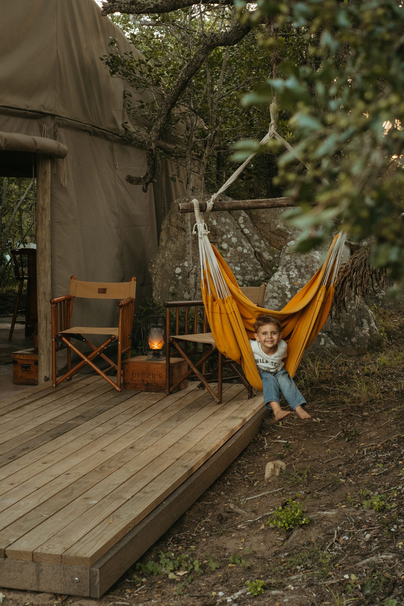 A boy grinning from a hammock chair beside a tent deck, a lantern on a crate