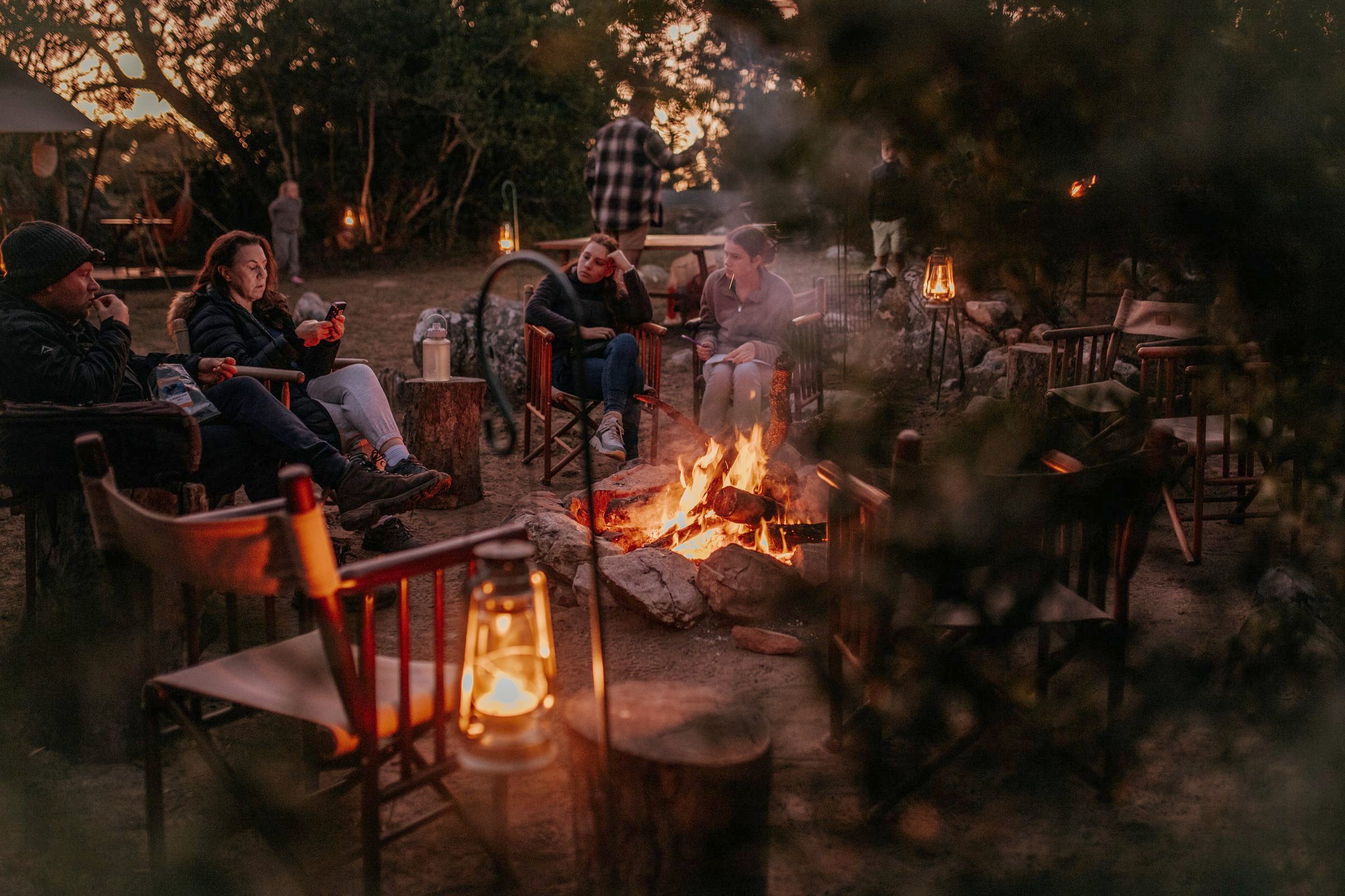 Guests in camp chairs around the fire circle at dusk, lanterns lit