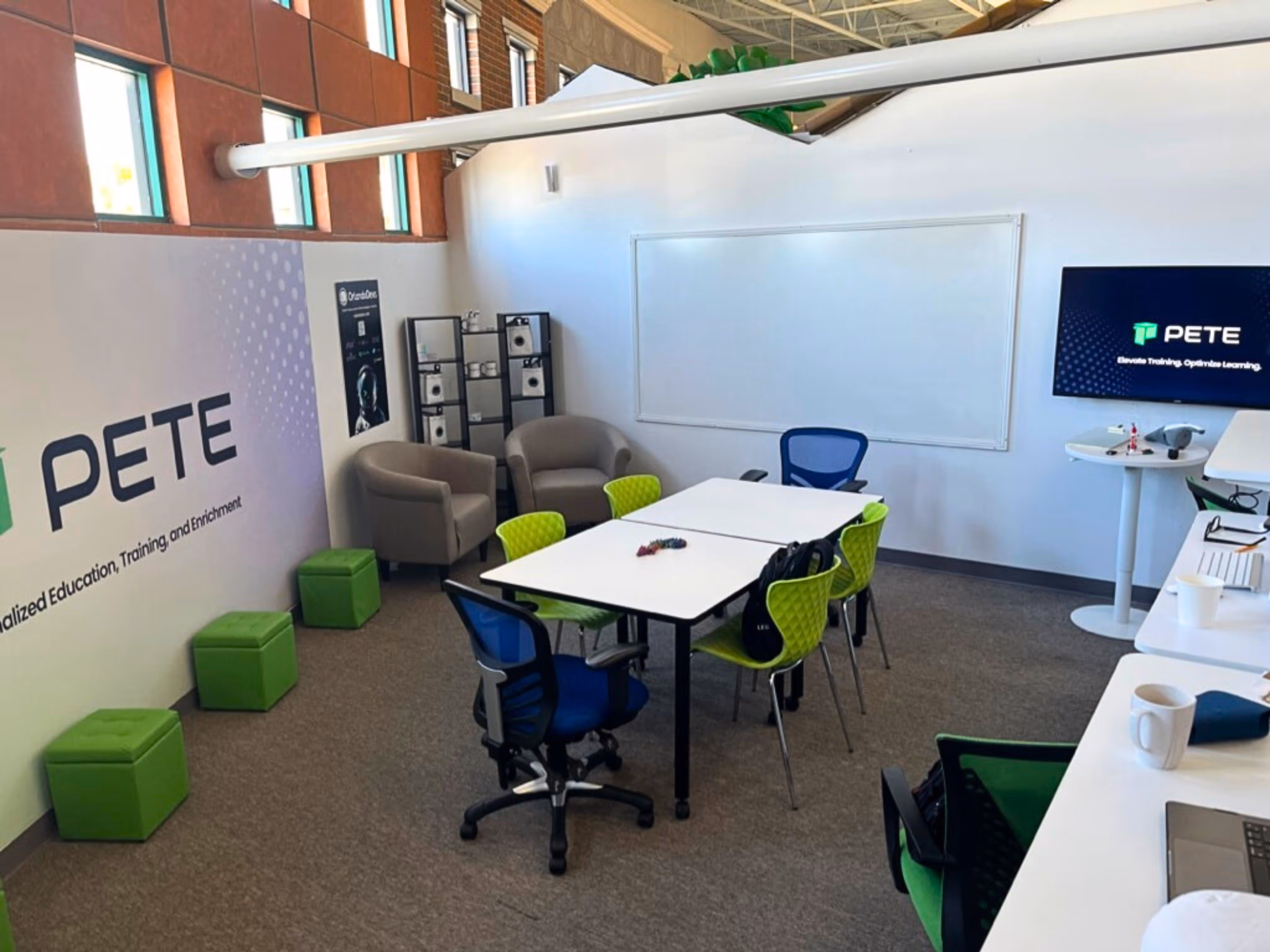 A picture of PETE's office space in Orlando, showing a wall panel with PETE's logo, a desk with PETE green-theme chairs, a drawing board and a TV screen mounted to the wall with PETE's logo.