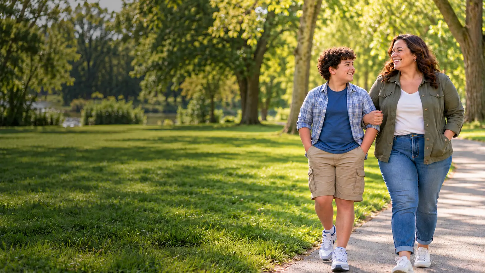 A smiling boy and his mom walking arm in arm through a sunny park