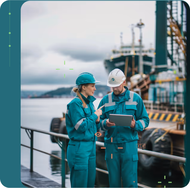 Two marine workers in green protective gear and helmets discussing while looking at a tablet near a docked ship.