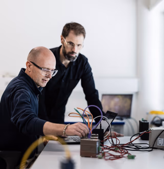 Two men working with electronic equipment and colorful wires on a desk in a bright room.