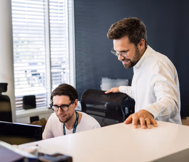 Two men wearing glasses working together in an office with computers and window blinds in the background.