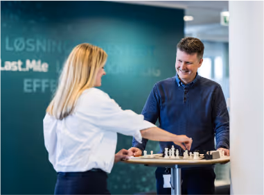A man and a woman play chess at a standing table in a modern office setting.