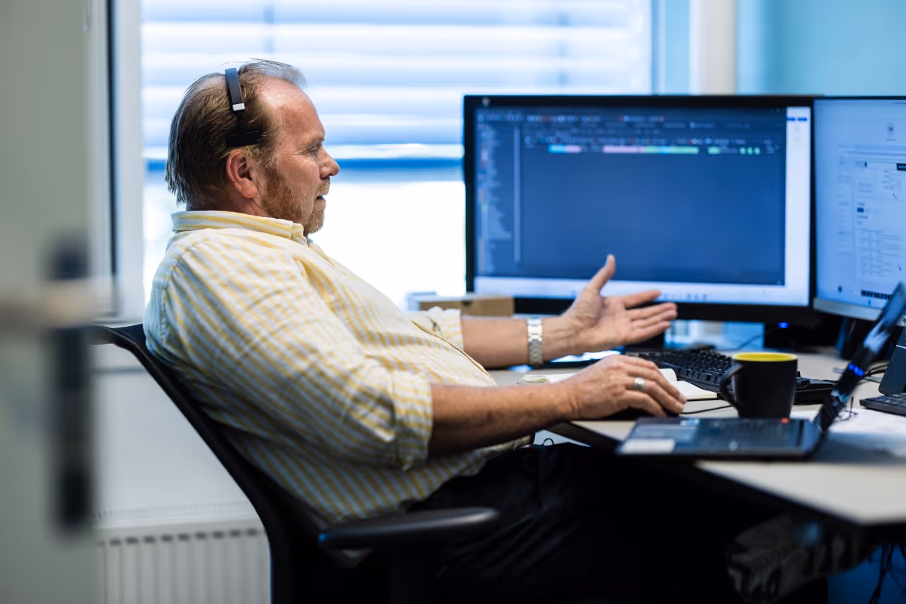 Man wearing headset and yellow striped shirt sits at desk using computer with dual monitors displaying software code and documents.