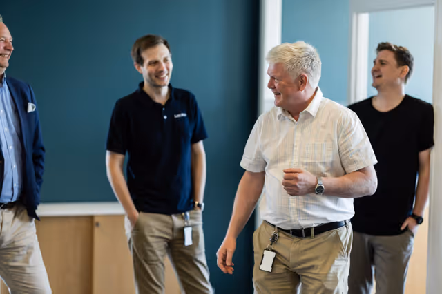 Four men casually talking and smiling in an office setting with blue walls.