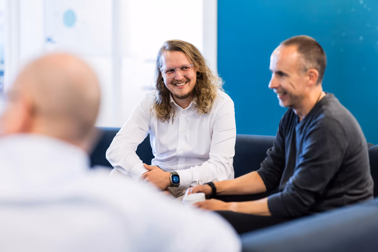 Three men sitting on a couch in an office, smiling and engaged in conversation.