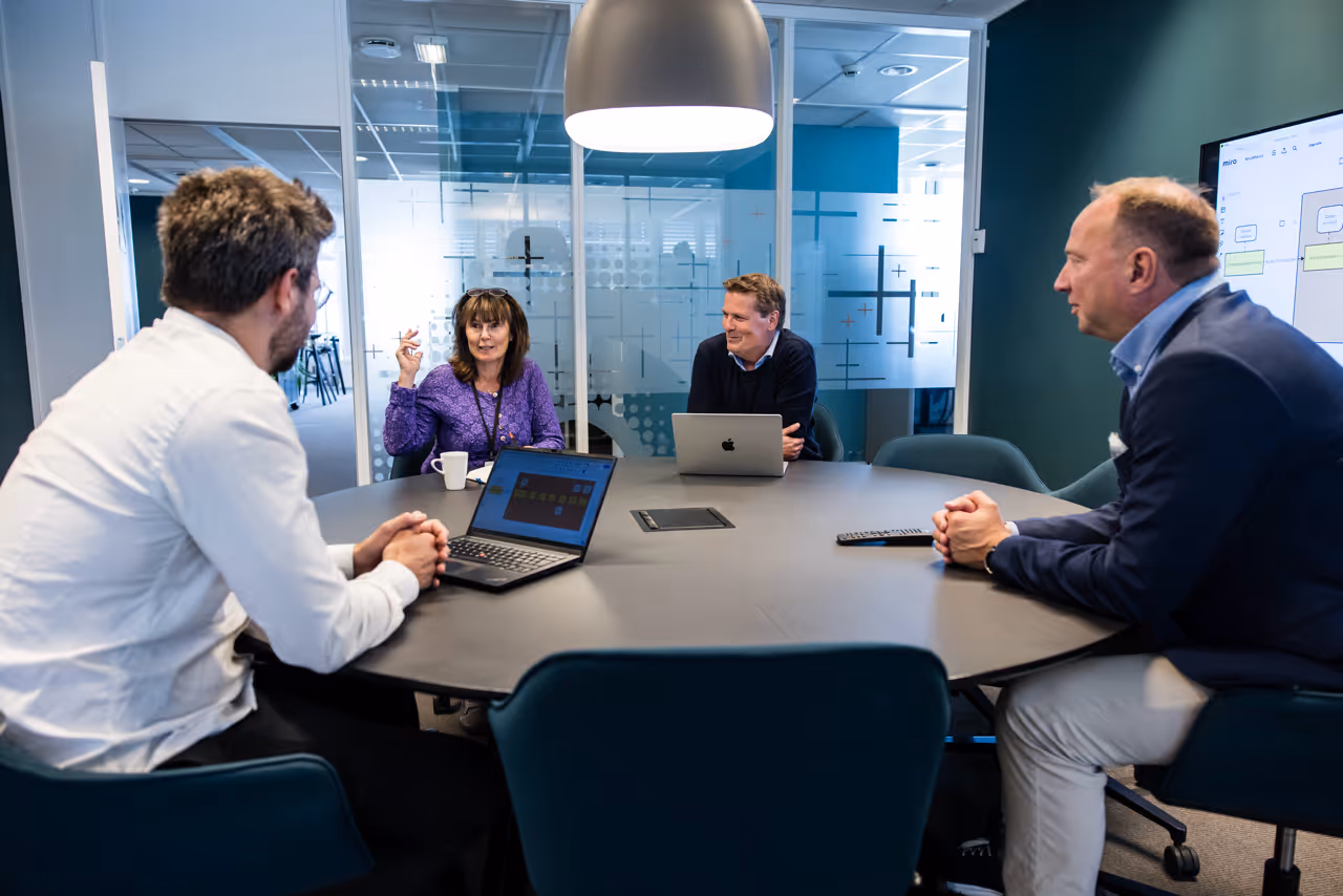 Four colleagues having a discussion around a round table in a modern office meeting room, each with laptops open.