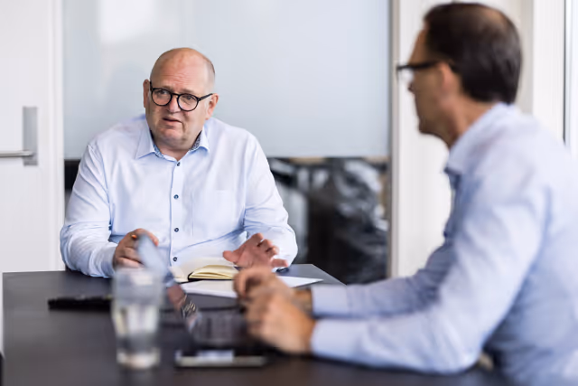 Two men in white shirts engaged in a discussion at a table with a notebook and glass of water.